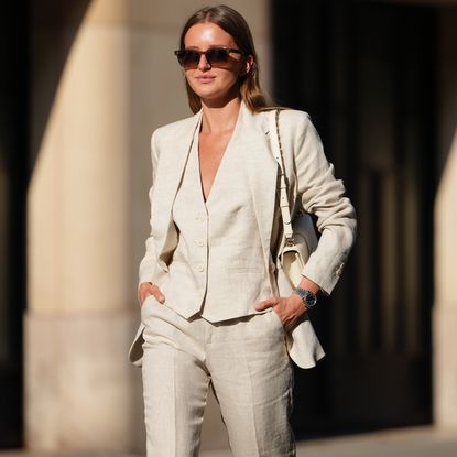Diane Batoukina wears a light beige linen suit consisting of a single-breasted blazer with notched lapels and matching trousers. The blazer features a tailored fit with buttoned cuffs. Underneath, a matching waistcoat with a V-neckline and button fastening is visible. Sunglasses with dark lenses and a tortoiseshell frame are worn. A white leather shoulder bag with a gold chain strap is carried. A metallic watch adorns the wrist. Hair is styled straight and parted in the middle, during a street style fashion photo session, on May 14, 2025 in Paris, France