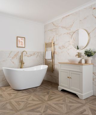 A white bathroom with a white and pink marble wall, a curved white freestanding bath tub with a gold tap, a white freestanding sink unit with a gold circular mirror above it, and brown parquet flooring