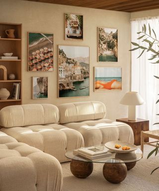 A living room with a large cushioned white couch, a beige wall with coastal art prints on it, and a glass coffee table with three circular legs