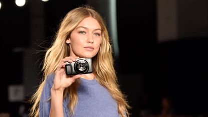 Gigi Hadid walking while pointing a compact camera at the person taking her picture.