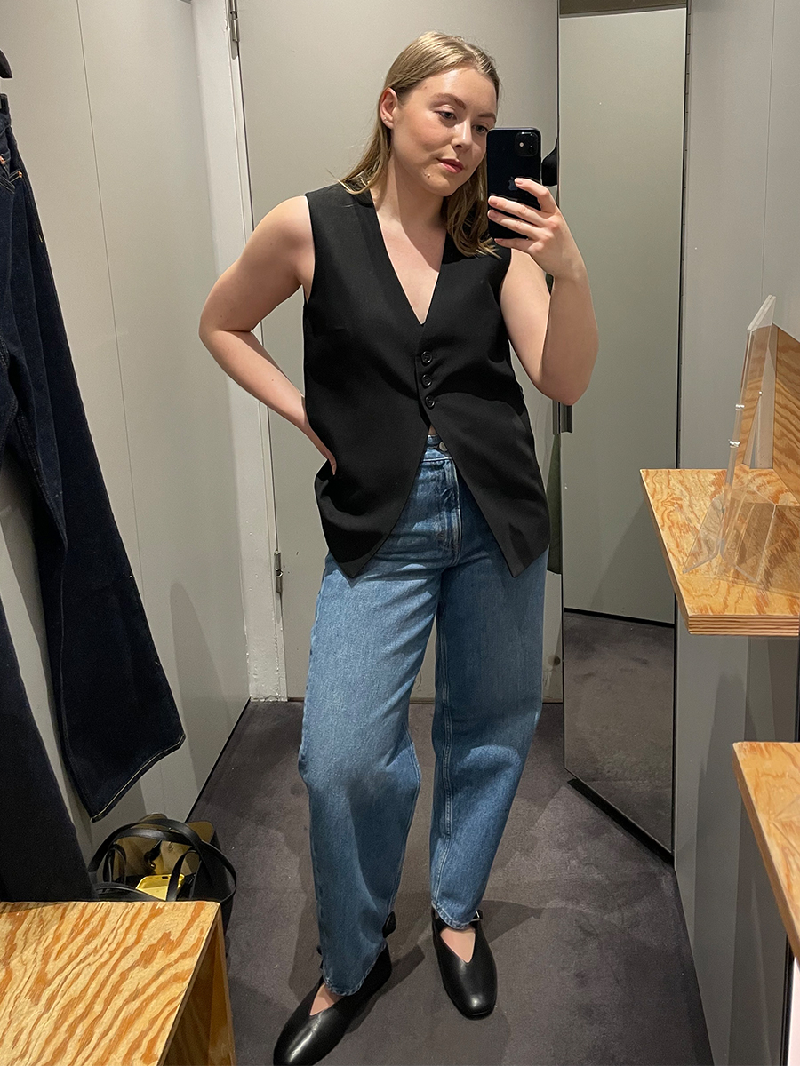 Woman in dressing room wears black waistcoat, blue jeans and mary jane flats