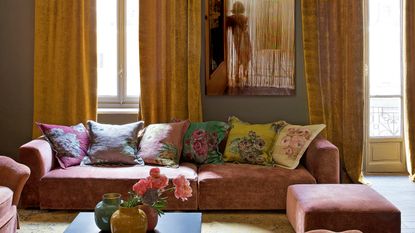 pink and ochre living room with ochre drapery, chandelier, pink modular sofa and chairs, colorful floral cushions, patterned rug and artwork of a lady in the