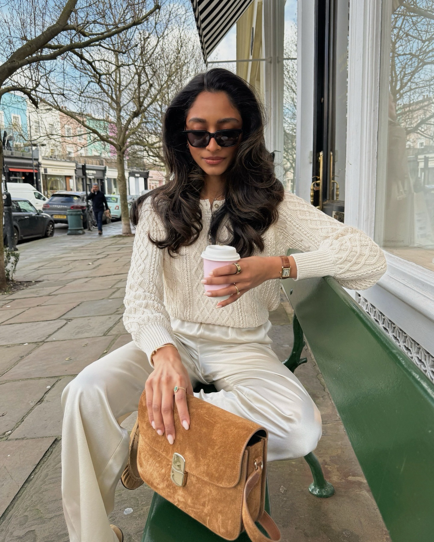 Fashion person Hannah wears a transitional winter to spring outfits 2026. She wears a white cable knit jumper, white satin trousers and butter yellow ruched ballet flats. She accessories with a suede crossbody bag from Sezane and black sunglasses and the Cartier Tank watch. She is sat on a green bench looking at her pink and white coffee cup in a recent image from her Instagram.
