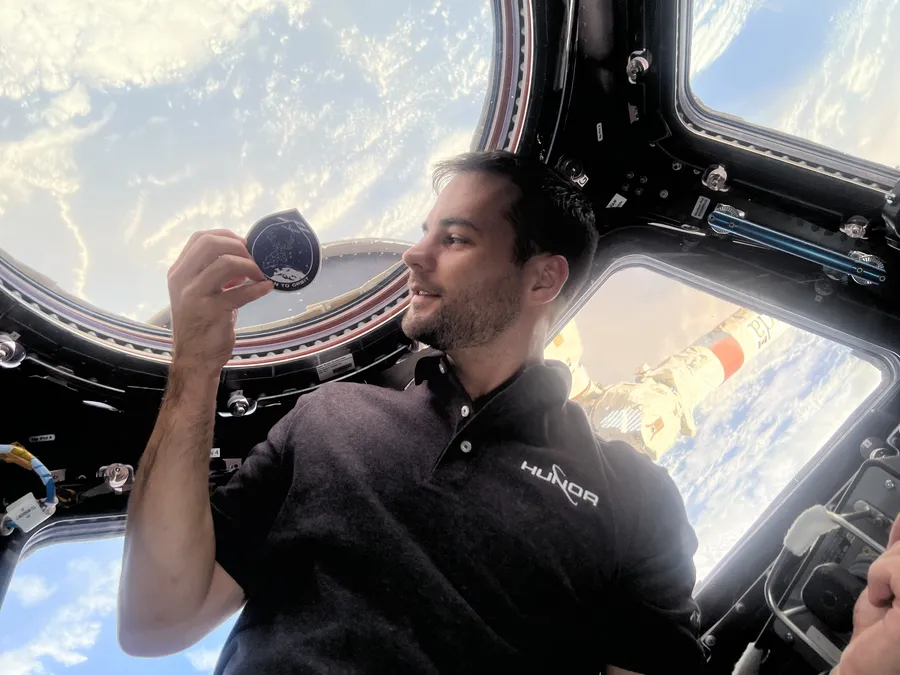 a man in a black polo floats in front of big windows in space that look down at a bright blue section of Earth.