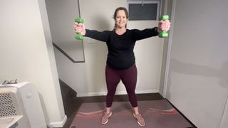 Certified trainer Jennifer Rizzuto performs an arms workout with dumbbells. She is standing with her arms out straight in front of her and a dumbbell clasped in each hand. She smiles at the camera; behind her we see a staircase and window.