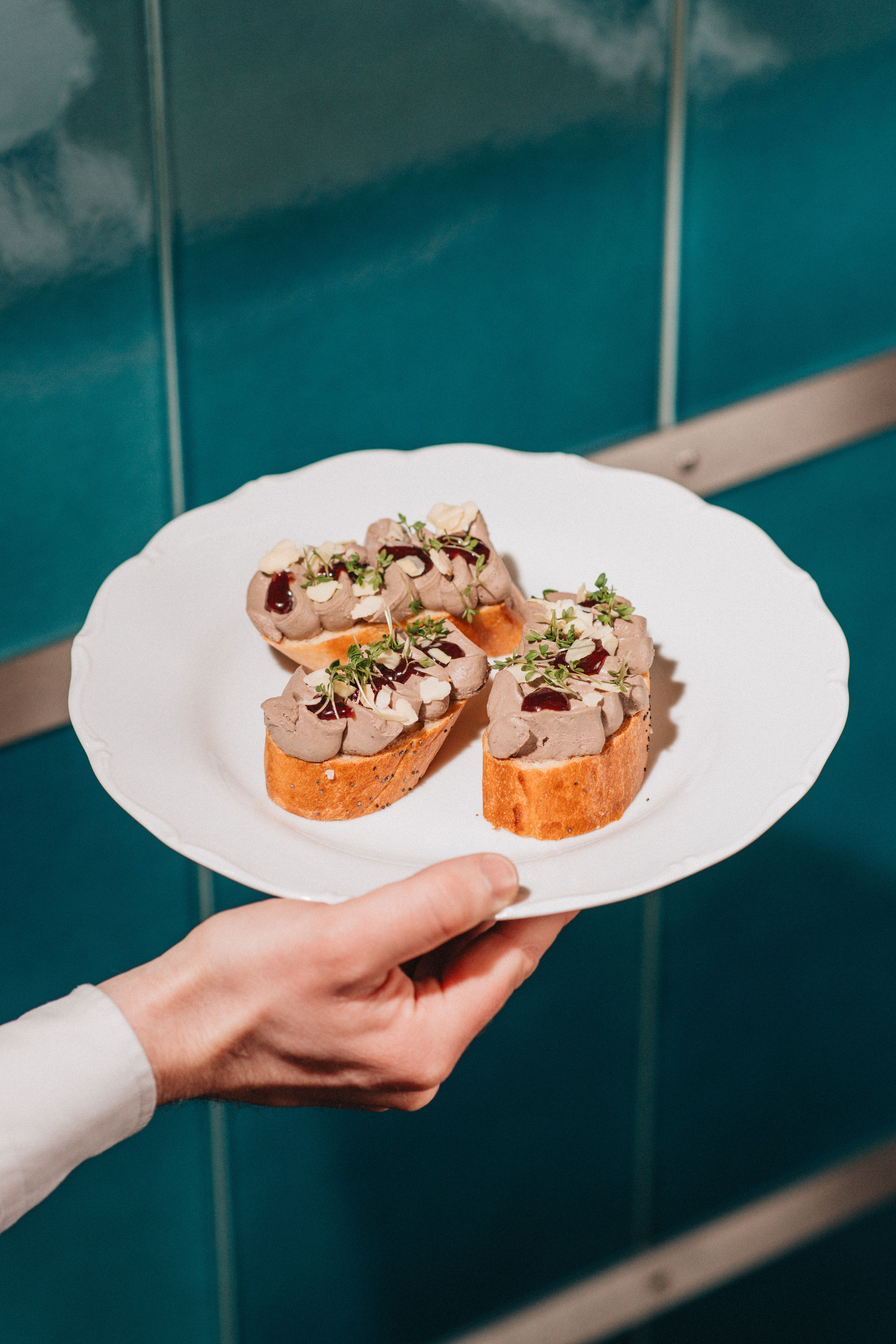 Two pictures of a restaurant, one caught from the inside of a sun-lit terrace, a waiter rushing by, and one of a plate of open sandwiches with fish, herbs, and olives, held by someone whose hand we see in front of a tiled water green wall.