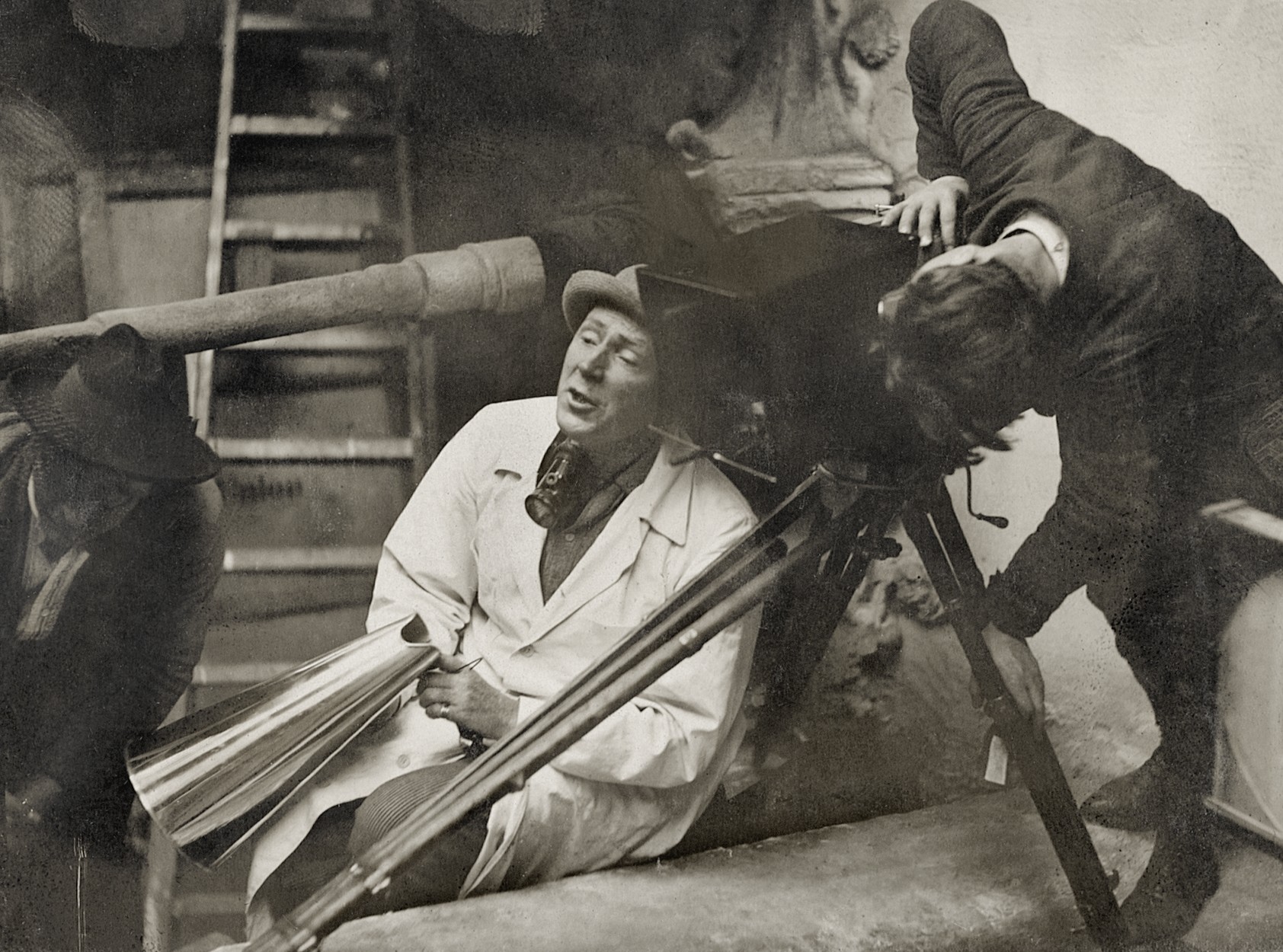 A man in a white coat and hat sits beside a large wooden film camera on a tripod, holding a metal megaphone, while another man peers through the camera&rsquo;s viewfinder.