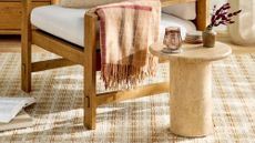 Joanna Gaines travertine table featured atop of a warm plaid-patterned area rug and mid-century-style wooden accent chair