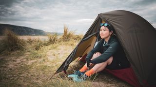 A woman sits in a MSR Hubba Hubba Bikepack One-Person Tent looking out, on a grassy, sandy beach.