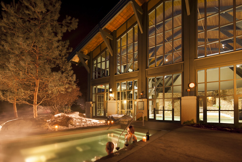 A couple is lounging in a hot-tub pool outdoors at nightime. An indoor pool in a wooden building with high ceilings is visible through large windows beyond. Courtesy of The Lodge at Woodloch.