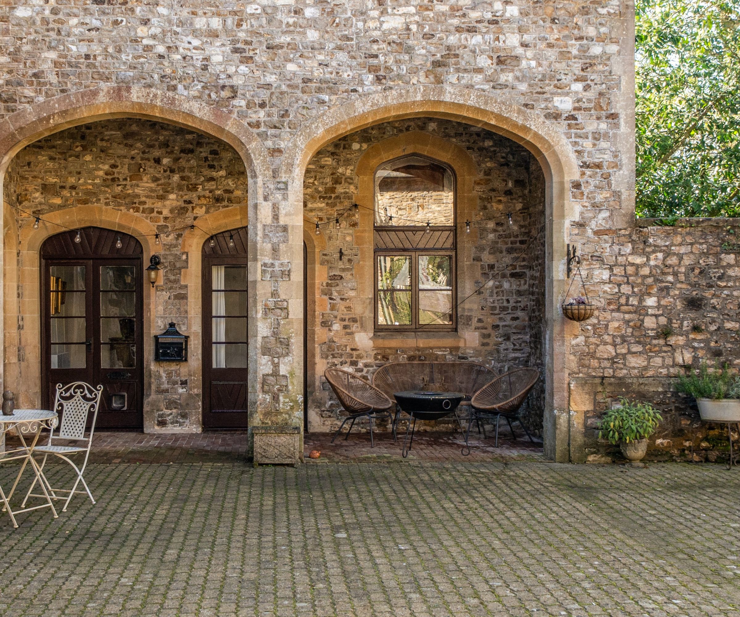 Stone-walled courtyard of a historic property with two arched bays, dark wood doors, a fire pit with wicker chairs, cobbled paving and fairy lights.