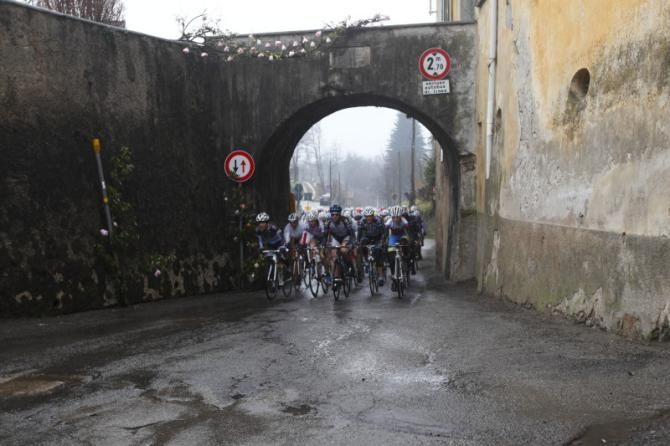 The peloton rolls beneath a bridge in a narrow village