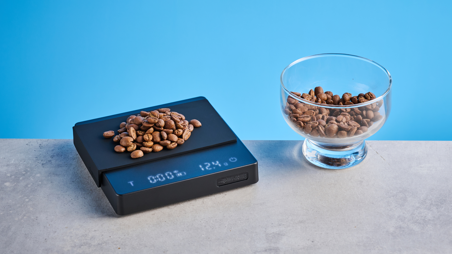 the mhw-3bomber cube coffee scale 2.0 in black photographed against a blue background showing its button controls and dial surface
