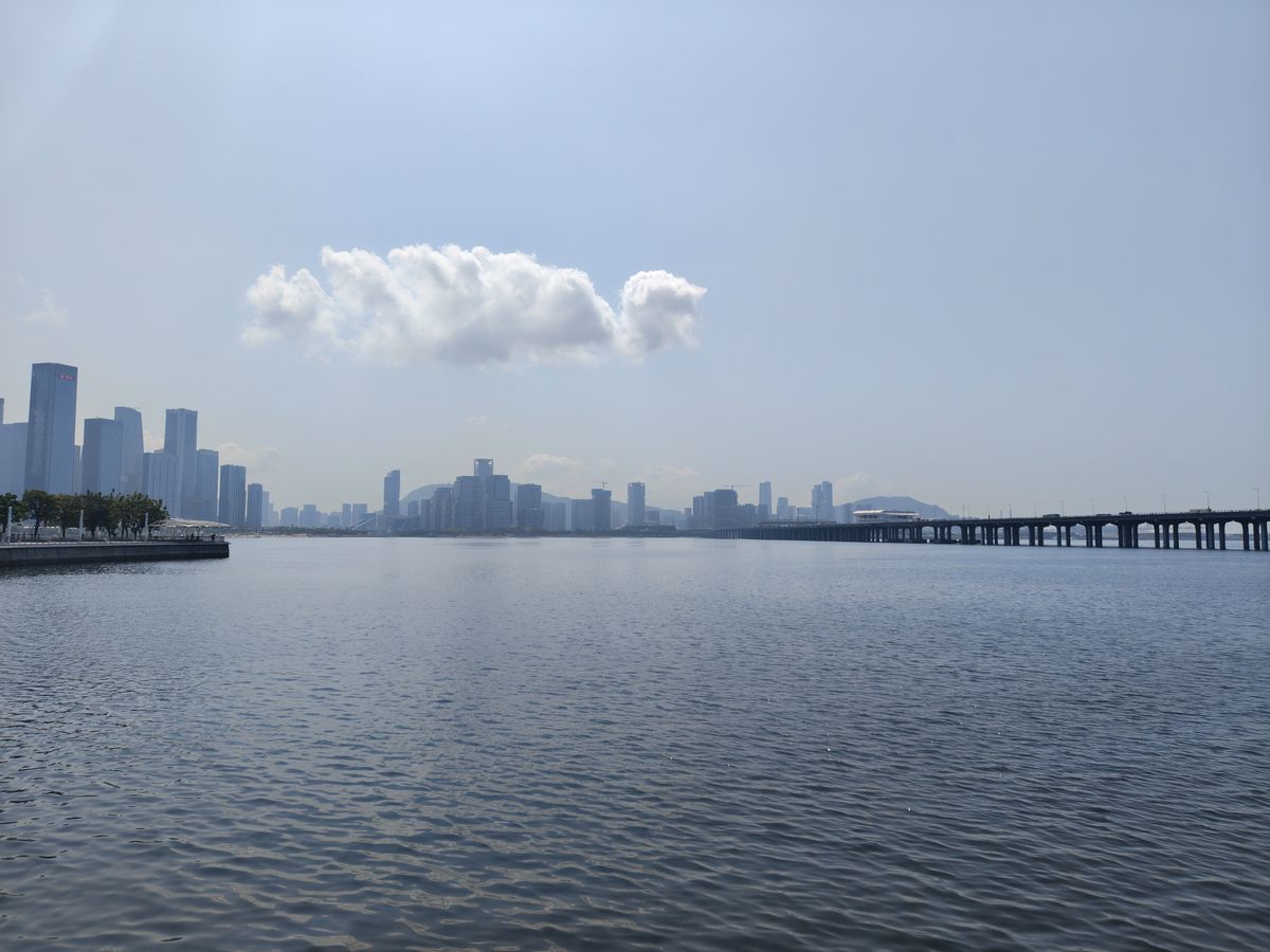 A wide landscape shot of a calm body of water with a city skyline in the distance. A long, low bridge spans the water on the right side of the frame. A single, prominent white cloud floats in the middle of a clear, pale blue sky, casting a soft reflection on the rippling water below.