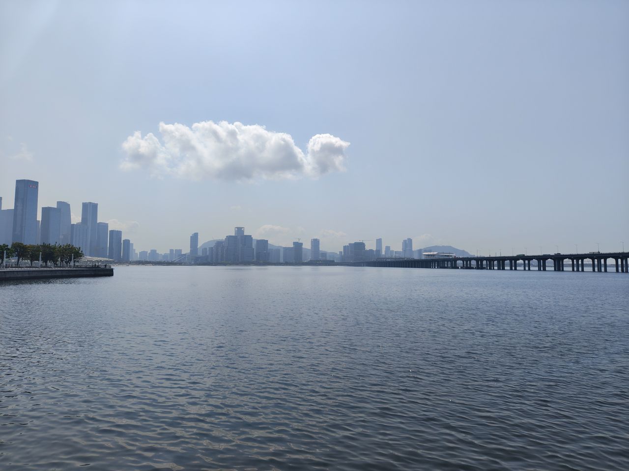 A wide landscape shot of a calm body of water with a city skyline in the distance. A long, low bridge spans the water on the right side of the frame. A single, prominent white cloud floats in the middle of a clear, pale blue sky, casting a soft reflection on the rippling water below.