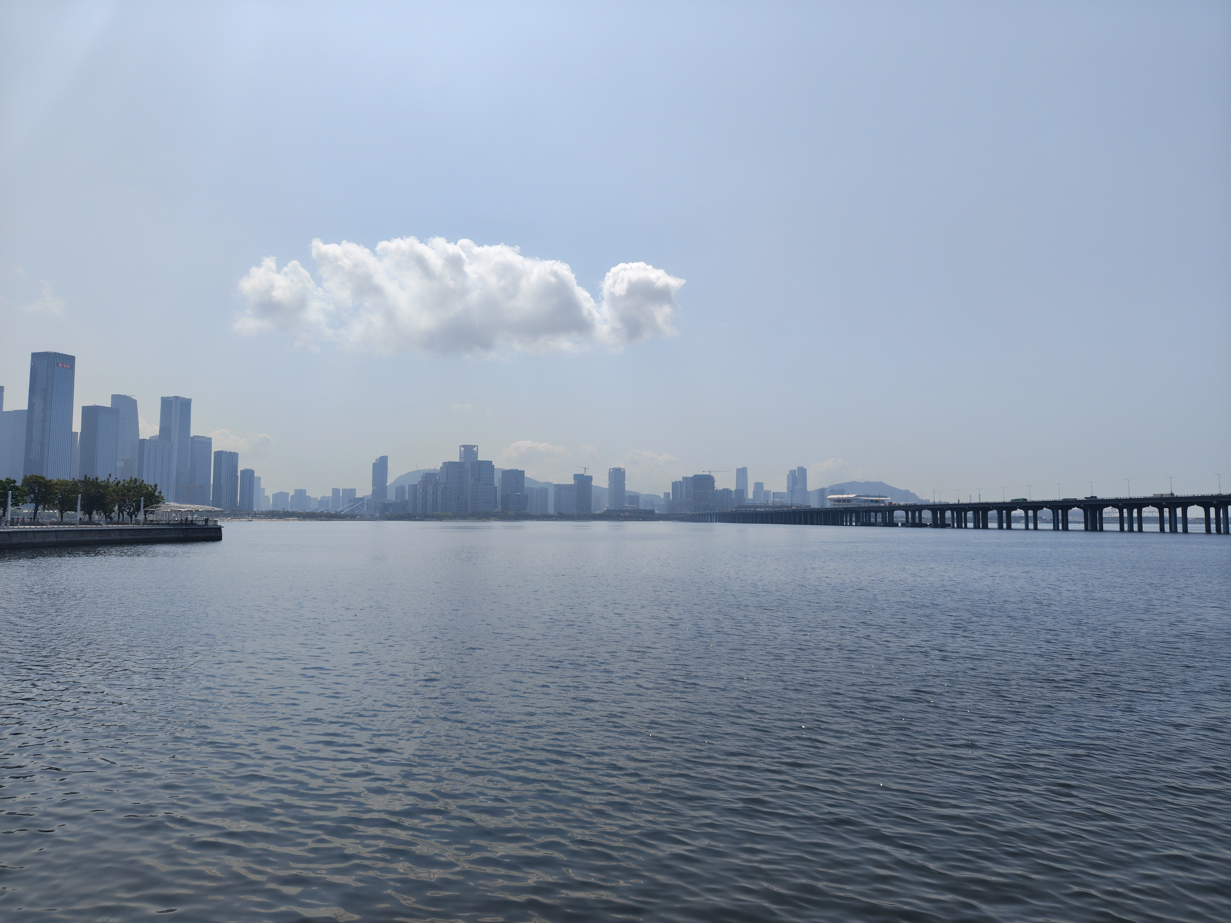 A wide landscape shot of a calm body of water with a city skyline in the distance. A long, low bridge spans the water on the right side of the frame. A single, prominent white cloud floats in the middle of a clear, pale blue sky, casting a soft reflection on the rippling water below.