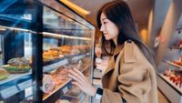A woman with straight dark hair wearing a smart watch and tan coat touches the glass on a bakery window, full of sandwiches.