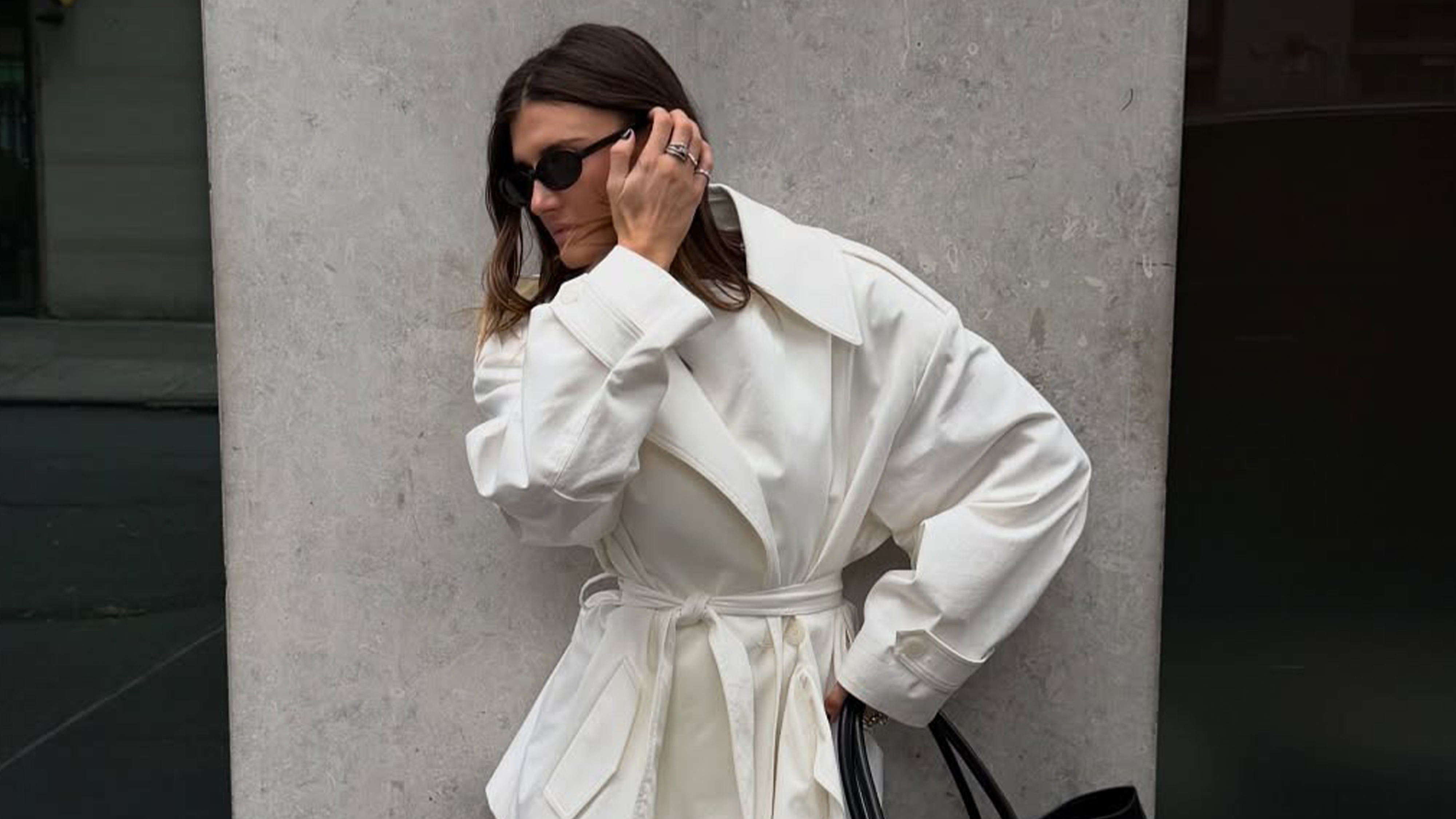 Influencer Coco Schiffer wears white coat, black bag, white socks and black loafers standing in front of New York wall.