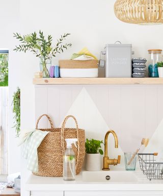 A neutral laundry room with wooden shelving, woven light fixture decor, woven baskets, grey laundry basket canister and wall paneling with geometric paint decor in white and grey with brass-colored faucet