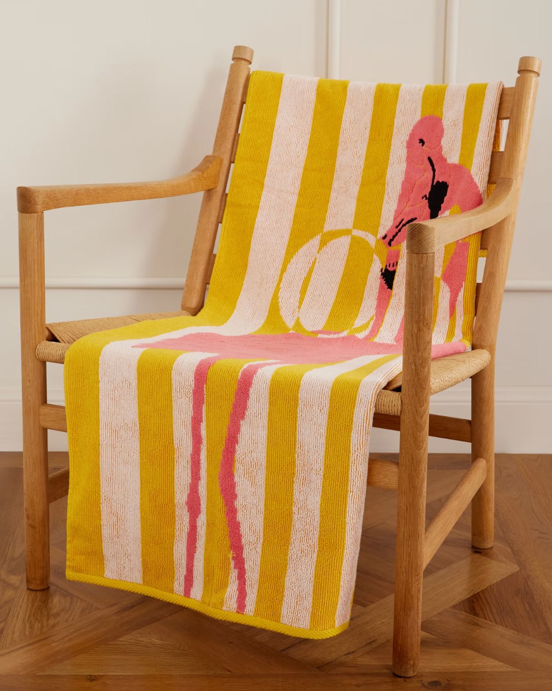 Cotton-Terry Beach Towel in yellow and white stripes, with a pink flamingo, open on a wooden beach chair in a wooden floored apartment.