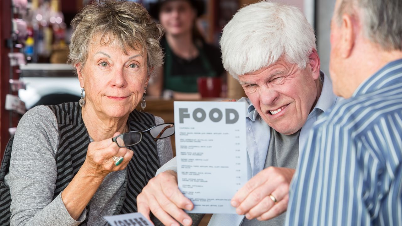 Older woman tries to hand her eyeglasses to an older man who&#039;s squinting at a restaurant menu.