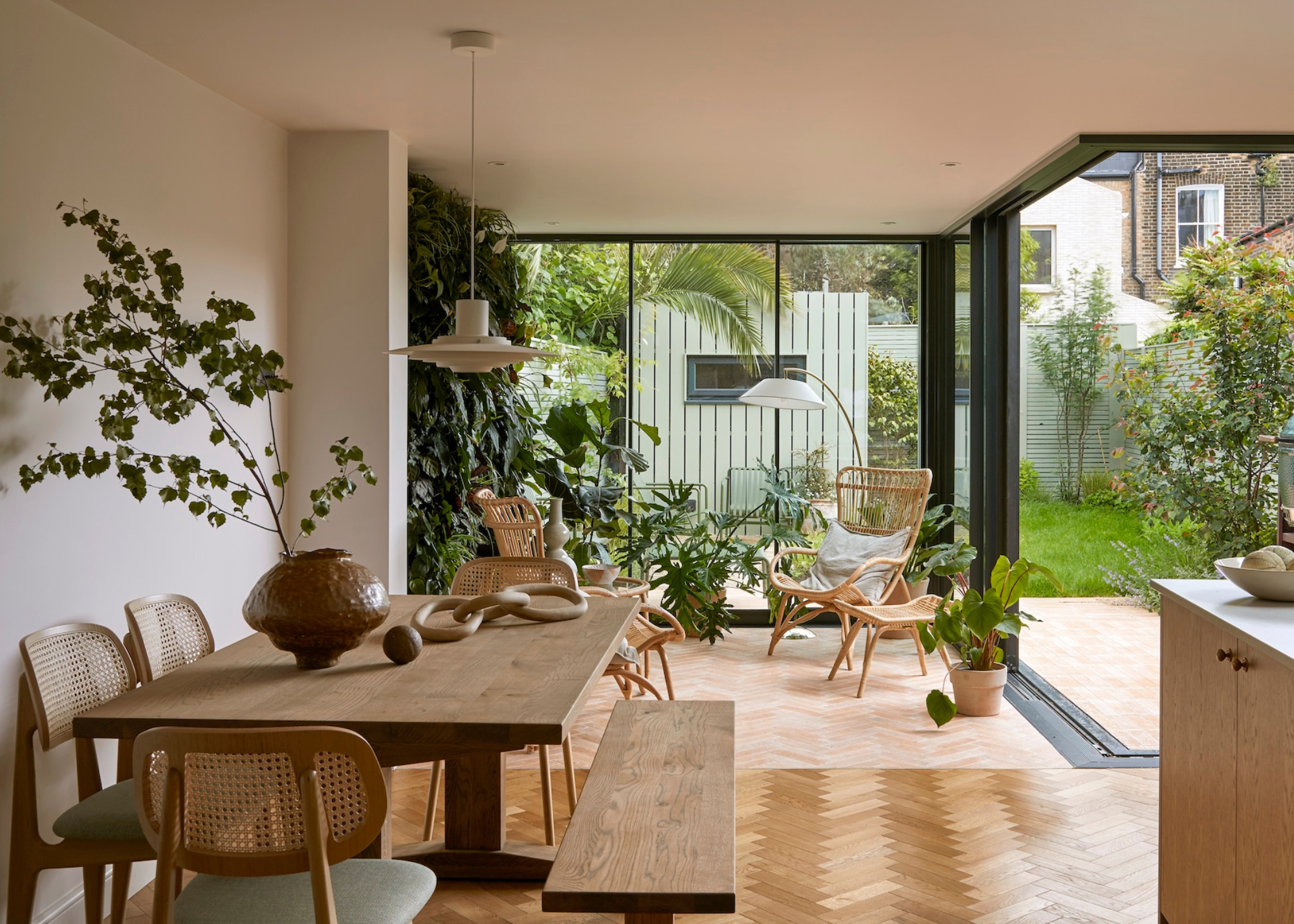 An indoor garden by a dining space with wood tables, rattan chairs, and woven armchairs leading to an outdoor garden