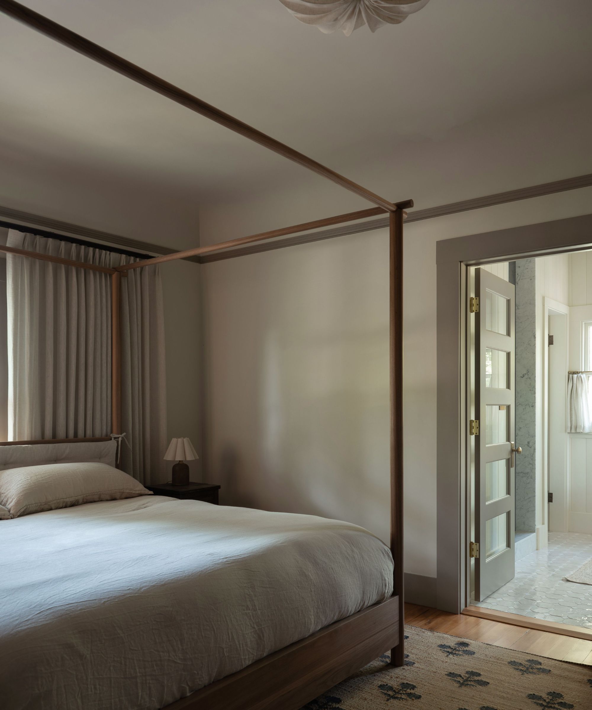 A modern, calming bedroom with taupe walls and dark mushroom trim, a minimal four-poster bed with crisp cream bedding and neutral curtains.