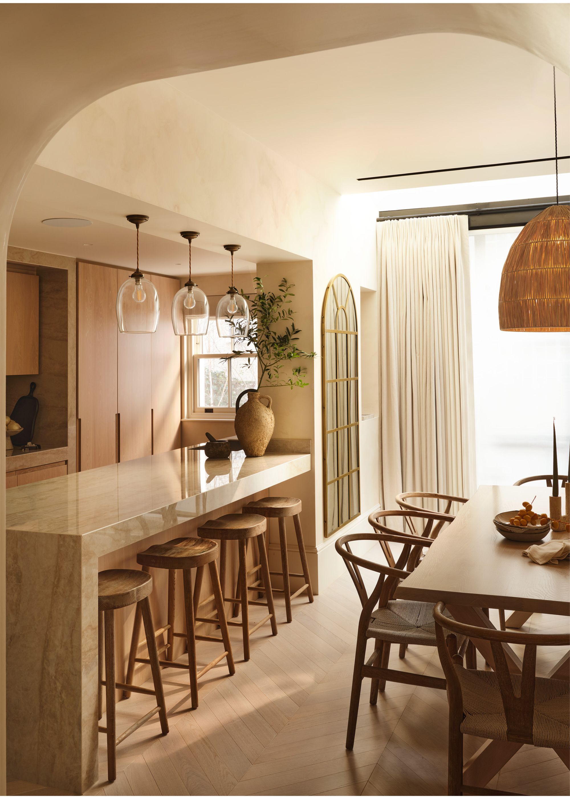 A modern kitchen in earth tones with a stone peninsula with three glass pendant lights above and four wooden stools tucked under the side closest to the dining table with curtains at the sliding door