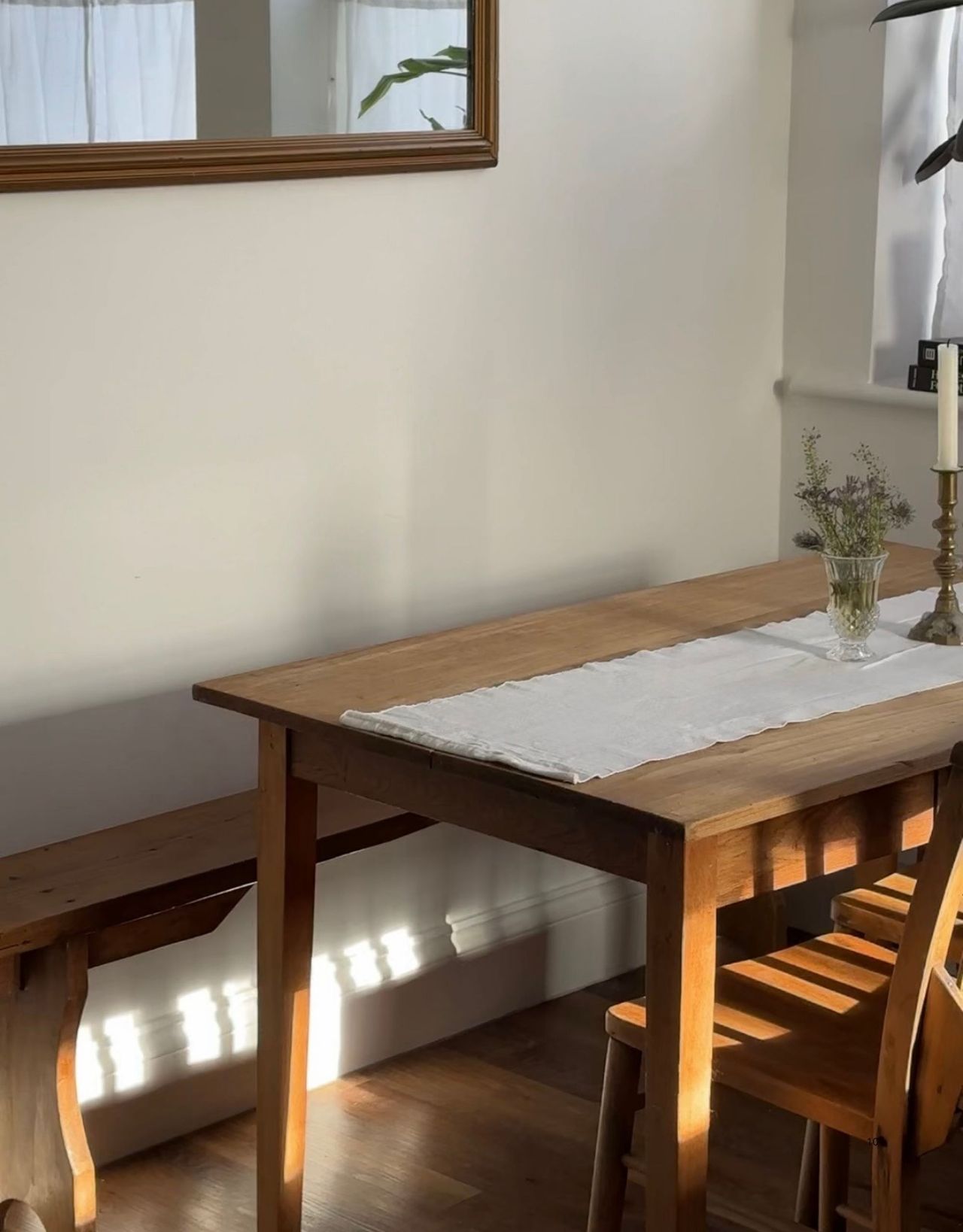 Image of a white breakfast nook with a wooden table and a vintage pew bench seat. There is a white runner on the table.