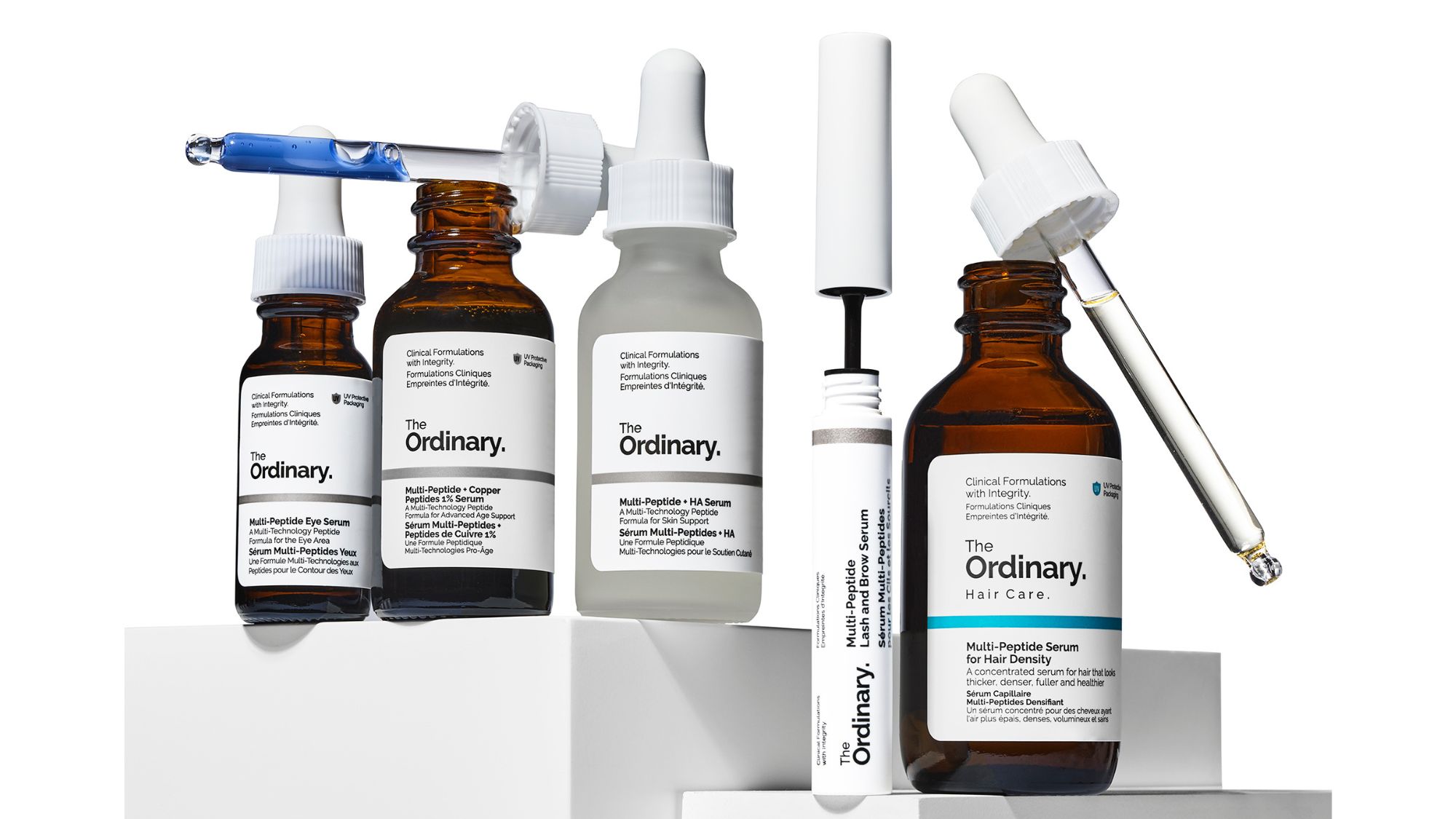 A selection of The Ordinary products lined up over a white background. Products include, from left to right: multi-peptide eye serum, multi peptide & copper peptides 1% serum, multi peptide + HA serum, multi peptide lash and brow serum, multi peptide serum for hair density