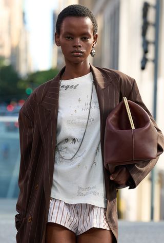 The frame bag trend is shown on the runway of Coach Spring 2025, with a model wearing a brown pinstripe blazer over a white graffiti print t-shirt styled with striped shorts and an oversized frame coin bag