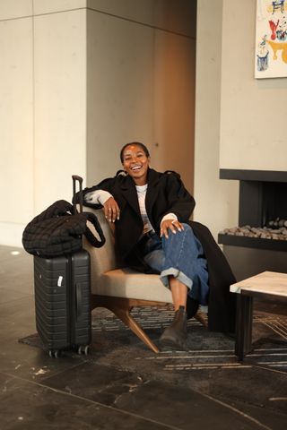 A woman sitting in a chair next to a suitcase and tote bag wearing a black coat and jeans