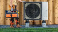 installer in work clothing with tool box stood next to heat pump installed outside timber clad property