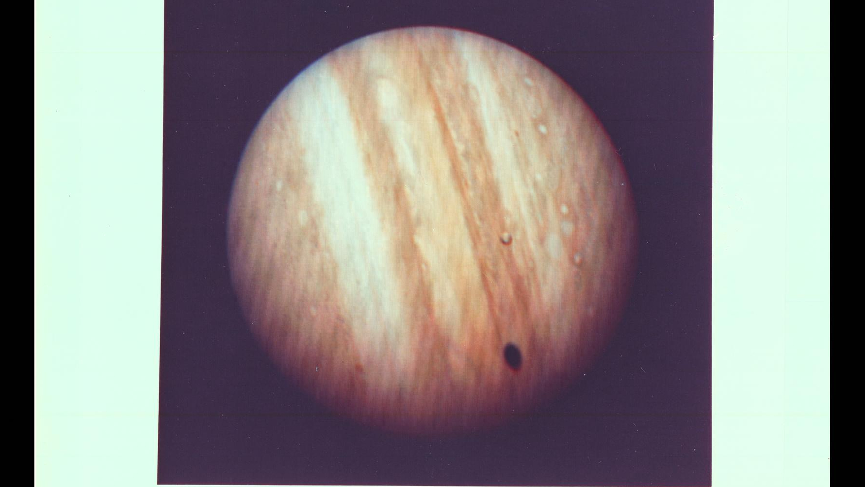 Jupiter imaged by Voyager 1 shows a brown/orange planet with a small moon in front. The shadow from Io is projected onto the Jovian atmosphere.