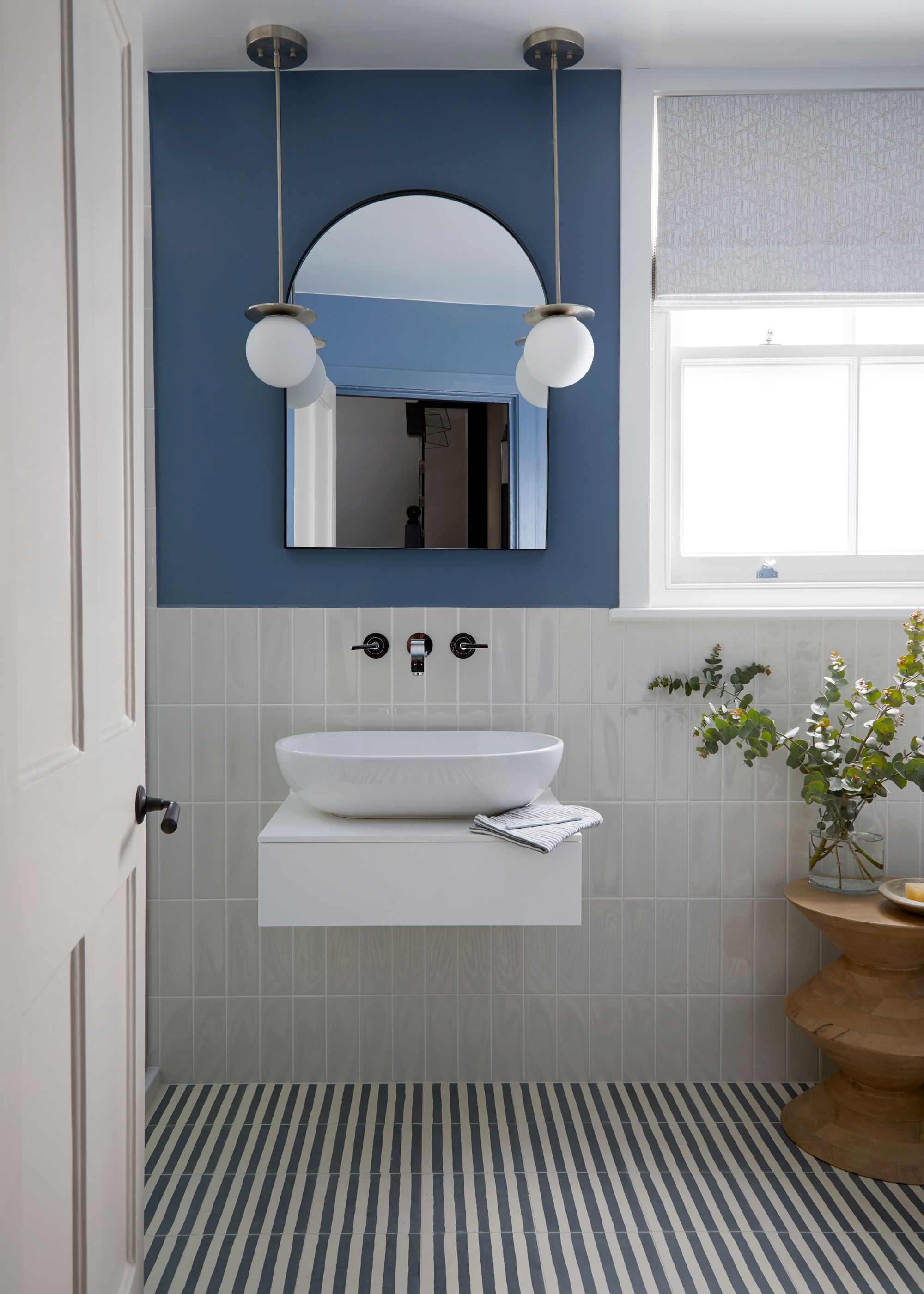 A modern bathroom with white tiles on the lower wall, a mid-tone blue paint on the upper walls, dark blue and white striped tiled flooring, a minimalist white basin sink, and an arched mirror above it.