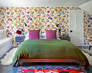 colorful bedroom with patterned wallpaper, bed with green duvet, pink cushions, white windowseat and blue bedside chests