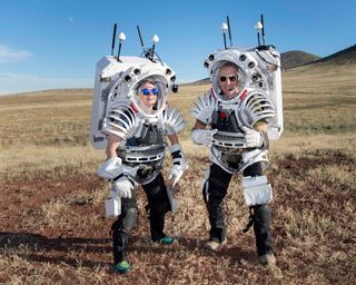 two nasa astronauts in a desert scrub field wearing mock white spacesuits with big antennas on top
