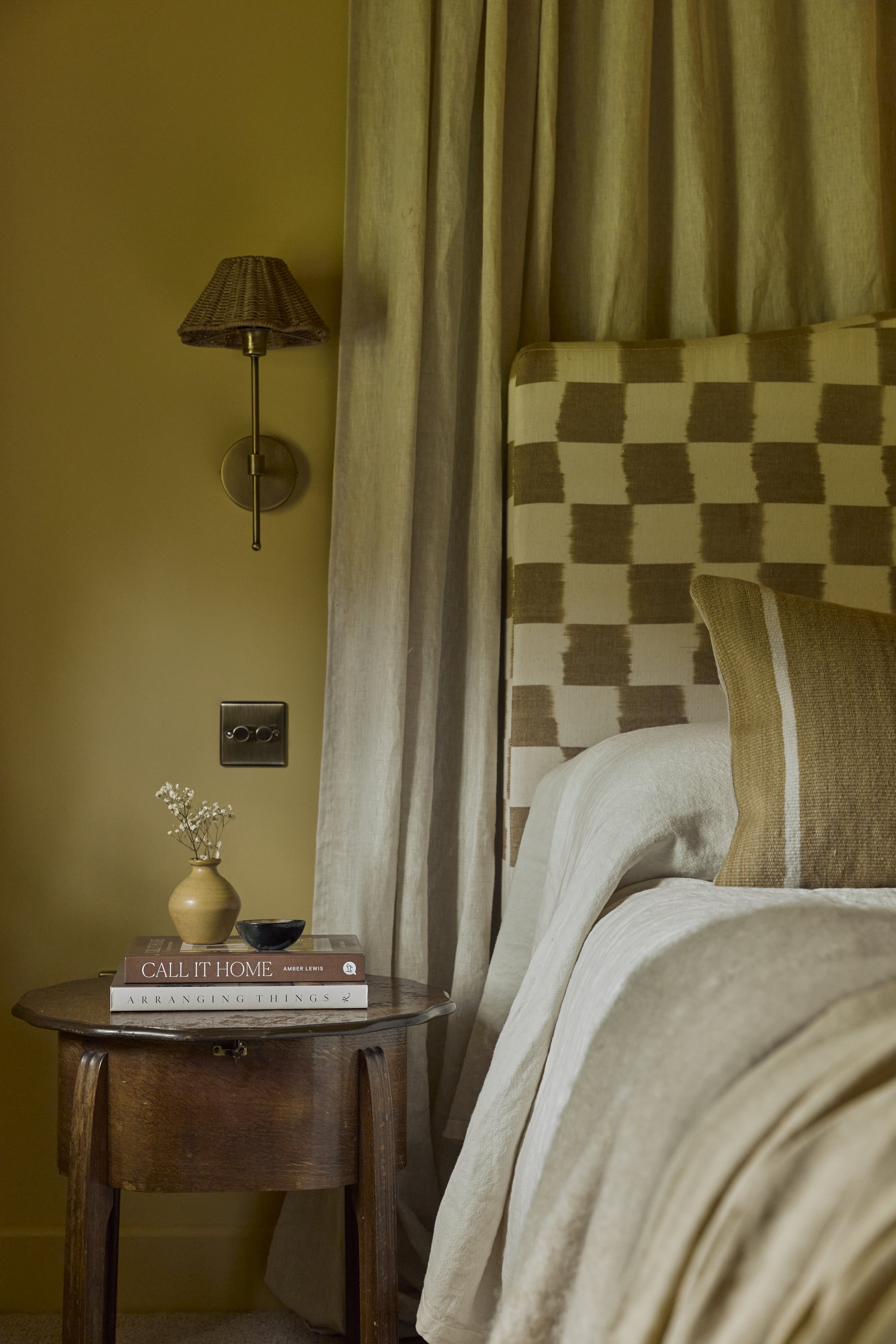 An ochre-hued bedroom features a series of matching furnishings, including a small ceramic pot filled with flowers, and striped and chequered bedding, along with wood and brass detailing.