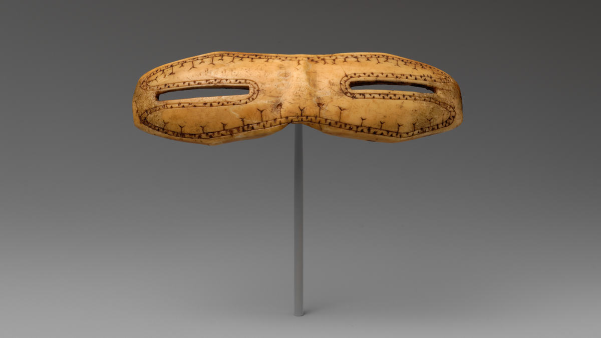 A carved piece of walrus bone in the shape of goggles with narrow slits at the eyes and incised decorations sits on a thin cylindrical stand against a grey background