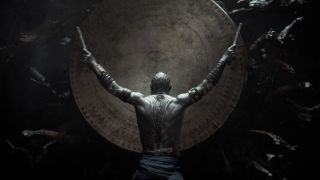 A gray human-looking alien is facing away from the camera towards a large drum. With both arms raised high, they have a thick drumstick in each hand. They have no shirt, but several bands/bracelets up both arms, a belt, and dark fabric trousers.
