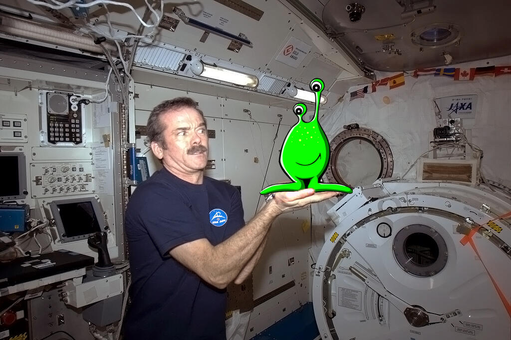 Chris Hadfield pictured on the left is holding what appears to be a small strange green alien with a big smile on it's face. He is holding the little green creature in the palm of his outstretched hands. Chris looks rather confused.