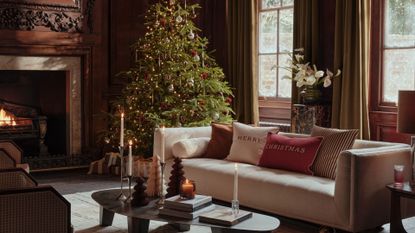 A living room with a Christmas tree, a white couch with festive cushions, a low coffee table with paper Christmas trees, taper candles, and stacks of coffee table books by a fireplace and a potted anthurium on a plinth