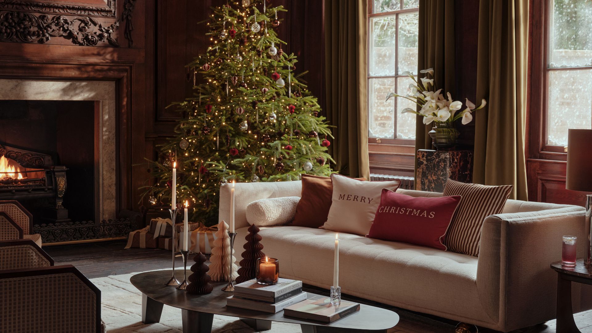 A living room with a Christmas tree, a white couch with festive cushions, a low coffee table with paper Christmas trees, taper candles, and stacks of coffee table books by a fireplace and a potted anthurium on a plinth