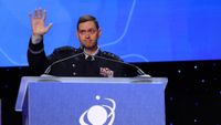 a man in a black military uniform with four silver stars on each shoulder waves while standing at a podium behind microphones