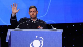 a man in a black military uniform with four silver stars on each shoulder waves while standing at a podium behind microphones