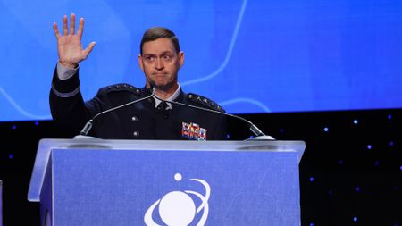 a man in a black military uniform with four silver stars on each shoulder waves while standing at a podium behind microphones