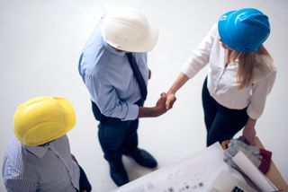 Three people in hard hats. A man in a shirt and tie shaking hands in agreement with a woman in a shirt with a blue hard hat