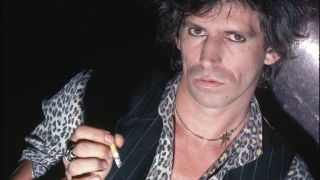 A close-up portrait of guitarist Keith Richards of The Rolling Stones. He wears a vest over a leopard-print shirt and holds a cigarette in his hand.