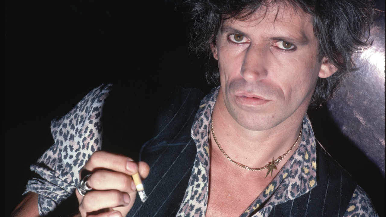 A close-up portrait of guitarist Keith Richards of The Rolling Stones. He wears a vest over a leopard-print shirt and holds a cigarette in his hand. 