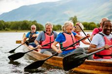 A group of older friends laugh as they row a long, wooden canoe.